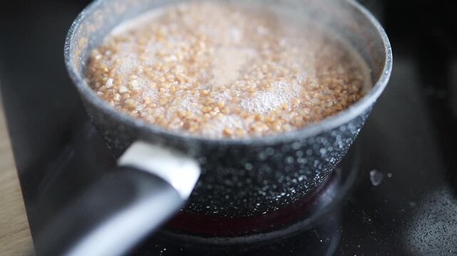 Buckwheat groats boiling and bubbling in a small saucepan on a kitchen stove during home cooking