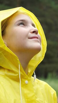 Profile view of a happy little girl with blue eyes in a yellow hooded raincoat looking up at the sky, enjoying the fresh air and feeling the rain on her face with a joyful smile