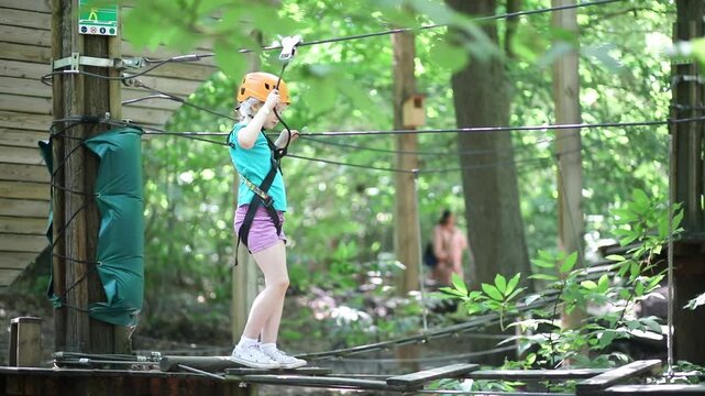 Young girl crossing an obstacle on a ropes course in an adventure park, wearing a helmet and safety harness among trees