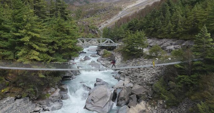 Annapurna Circuit, Manang region, Nepal, aerial glide shot traversing a deep valley to frame a traditional suspension bridge spanning the rocky banks of the Marsyangdi River.