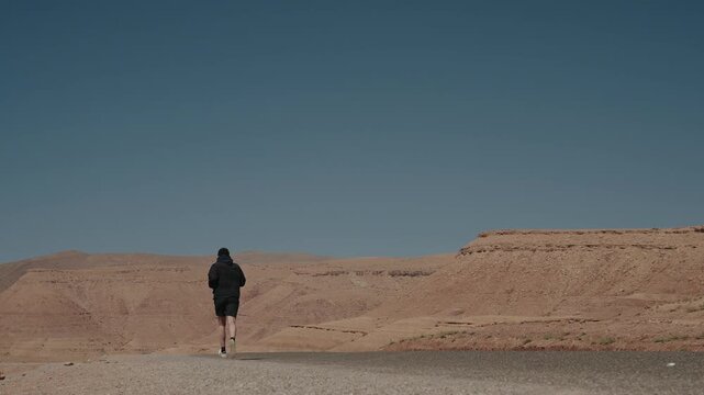 Runner in black hoodie jogs toward vast arid plateau under clear sky in Morocco desert