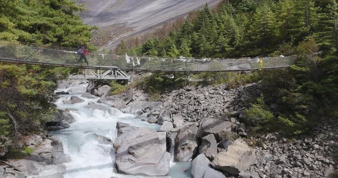 Annapurna Circuit, Manang region, Nepal, aerial gliding towards Swargadwari peak over rocky waters of the Marsyangdi River, steel truss footbridge, a trekker walking on the trail toward pine forest,