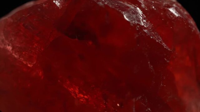 Extreme macro close-up of a raw, rough red crystal or mineral specimen, displaying jagged natural facets and translucent deep ruby color against a dark background.