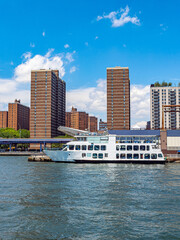 Fototapeta premium White passenger ferry docked beside New York apartment towers. Broadside view shows two decks, rows of windows and people at an East River pier under blue sky.