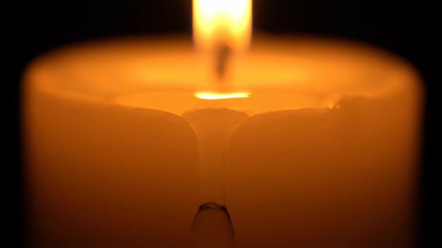 Warm glowing candle flame melting wax and forming a large drop against a deep black background, symbolizing hope, memory, or peaceful ambiance.
