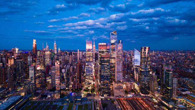 Hudson Yards skyline under layered clouds at blue hour. Aerial view shows 30 Hudson Yards, the Vessel, rail yards and illuminated glass towers beneath textured clouds.