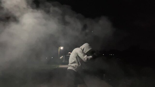 Cinematic slow motion of a hooded man practicing shadow boxing in dense smoke under a street light at night.