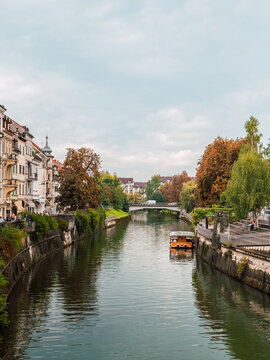 Scenic view of the Ljubljanica River winding through Ljubljana, with historic riverside buildings, lush trees and a tourist boat under cloudy skies in Slovenia