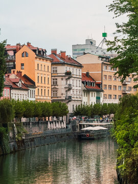Colorful historic buildings lining the Ljubljanica River in Ljubljana, Slovenia, with calm reflections, riverside promenade and urban architecture under an overcast sky