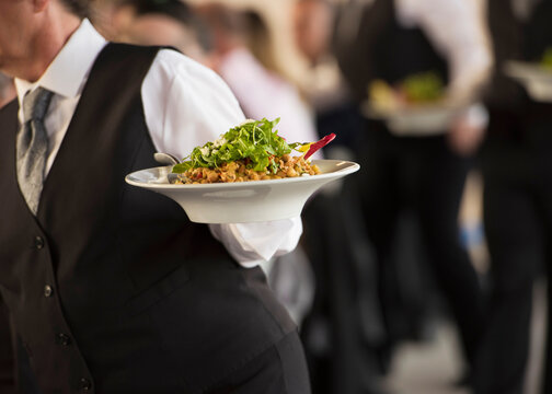 Banquet server carrying a plated salad during formal event service while fellow waitstaff follow behind, illustrating hospitality, catering, teamwork, and professional food service with faces unseen.