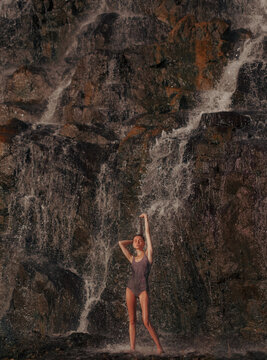 Beautiful woman in swimsuit standing under waterfall and splashing water