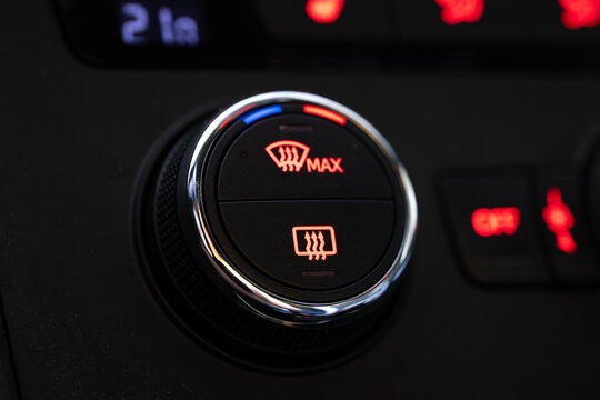 Close up of a car dashboard climate control dial with illuminated red windshield defroster symbols and chrome ring.