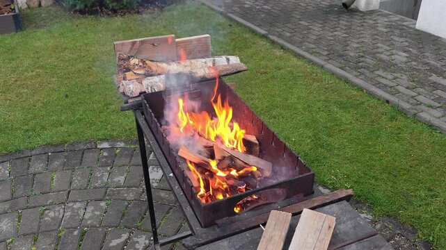 On a tiled area near a grassy lawn there is a metal brazier in which wood burns and crackles and smoke comes out. There are birch firewood nearby. Overcast autumn weather