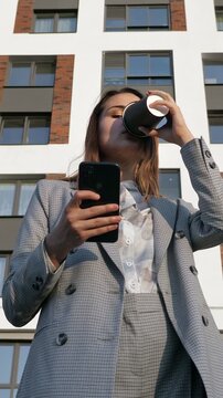 Young businesswoman or realtor in a formal suit checking her phone and drinking coffee with a modern residential building in the background, embodying a successful urban lifestyle