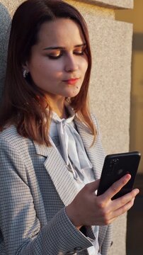 Beautiful young professional woman enjoying a quiet moment outdoors, smiling as she types and browses on her mobile phone, illuminated by the beautiful golden hour light