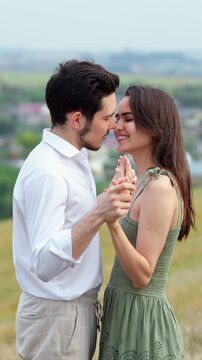 Beautiful young couple in love holding hands, smiling, and looking at each other with affection while standing close together on a hill, enjoying a romantic date outdoors in nature
