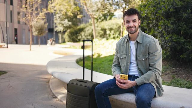 Portrait of man with suitcase sitting park on bench using smartphone smiling to camera
