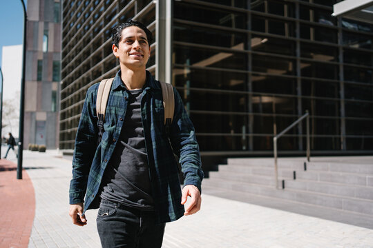 Young hispanic student walking outdoors near modern office university building in urban city carrying backpack