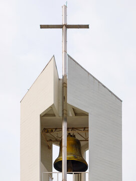 A low angle shot of a modern architectural church bell tower featuring a large brass bell and a Christian cross on top against a bright sky background, religion and architecture concept.