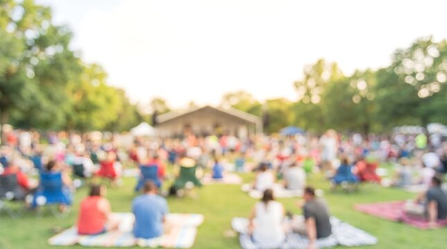 People sitting on lawn chairs at outdoor concert in park with blurred audience and stage background during summer evening music festival