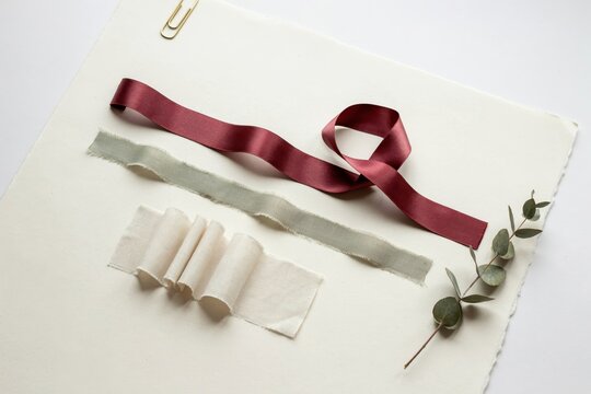 Flat lay composition of burgundy silk ribbon, sage green frayed fabric, and eucalyptus sprig on textured off white paper with gold clip