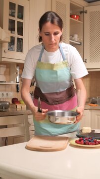 vertical video Woman in an apron is carefully weighing ingredients on a kitchen scale, preparing a healthy meal with fresh berries and cottage cheese, focusing on a balanced diet and home cooking