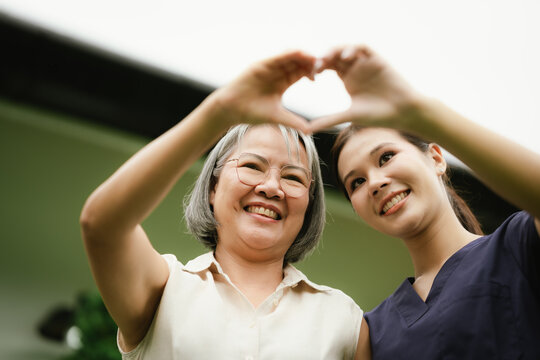 Smiling senior woman and caregiver making a heart shape with their hands, expressing love, trust, compassion, and quality elderly care.