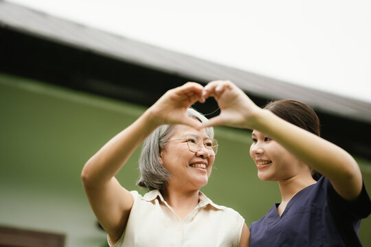 Smiling senior woman and caregiver making a heart shape with their hands, expressing love, trust, compassion, and quality elderly care.