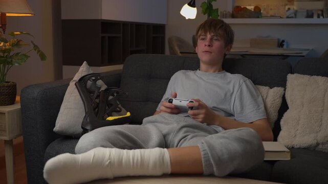 Teenager with cast on broken leg getting upset and throwing controller after losing video game, recovering from snowboarding injury on comfortable sofa in living room at night