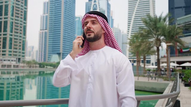 Arab man with a trimmed beard wearing a traditional red and white checkered keffiyeh and a white kandura, talking on a smartphone