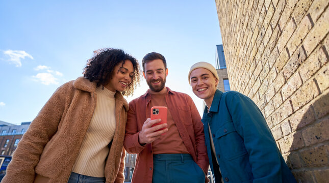 Urban friends looking at smartphone in autumn coats against brick wall