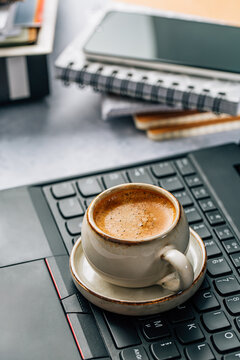 A cup of espresso sits on a laptop keyboard next to notebooks and a phone