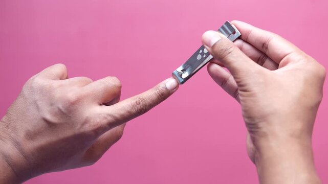 Hand using nail clippers to trim fingernail on pink background, close up detail shot