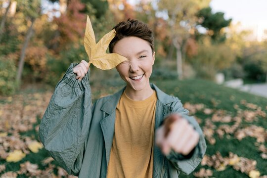 Vertical portrait of a smiling teen holding a yellow maple leaf over one eye and pointing at camera in an autumn park for seasonal banner and mobile wallpaper