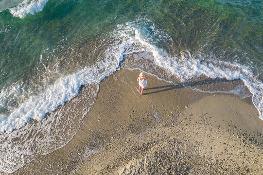 A woman in a white dress and straw hat stands on the shore of a sandy beach. The ocean's surface washes over her feet. View from above, from a drone.