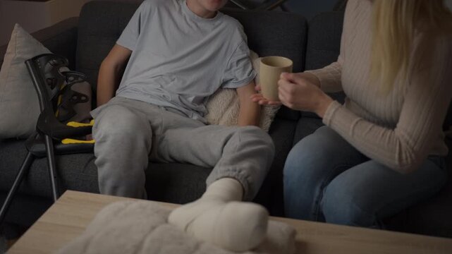 Caring mother giving warm drink to her teenage son with a broken leg resting on the couch, showing family support, love, and care during his injury recovery at home