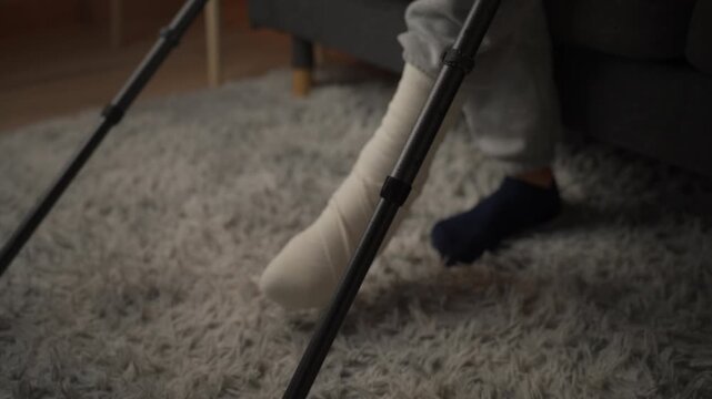 Close-up view of a person with a bandaged leg after an injury, carefully standing up from a sofa and beginning to walk with the aid of crutches, illustrating the recovery process