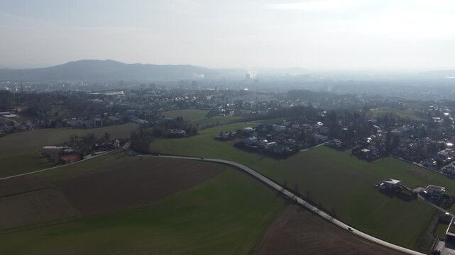 Aerial drone view over farmland and residential neighborhoods near Leonding, Austria, with a winding road and the hazy skyline of Linz, its industrial plants, and distant hills in the background