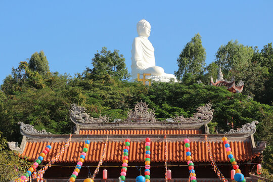 Horizontal panoramic view of the giant White Buddha statue on a hill rising above the traditional roof of Long Son Pagoda.