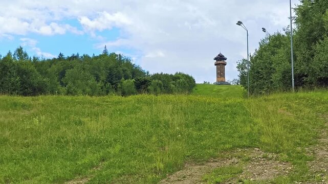 Chairlift tower made of steel framework clad with wooden planks, standing on a mountain slope in Gura Humorului against a mixed forest.