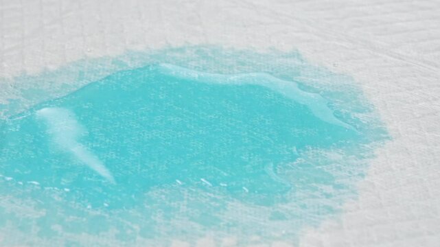 Blue liquid being absorbed by white disposable incontinence pad, close-up demonstration of absorbency performance