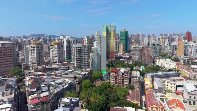 Parallax flight over dense Macau residential towers reveals crowded rooftops, green treetops, a turquoise church spire and hazy hills beyond