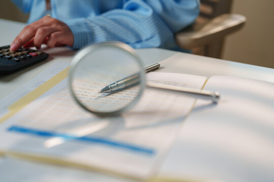 Close-up of financial documents being reviewed with a magnifying glass and calculator, auditing, compliance, financial analysis, and business verification in a professional office environment