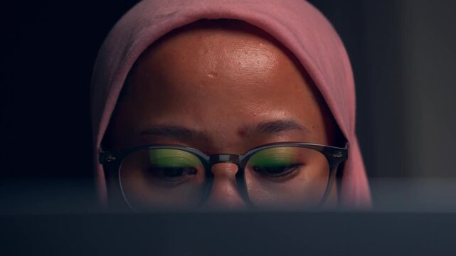 muslim woman with pink hijab and glasses stares at screen, working late concept