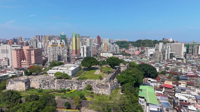Parallax aerial reveals stone walls of Mount Fortress and Macau Museum courtyard with tourists strolling green lawn against dense city skyline