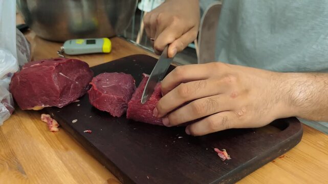 Male hands cutting raw beef into smaller pieces on cutting board before grinding meat in home kitchen, 4K.