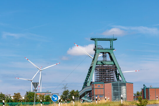 Old industrial mining headframe tower standing next to modern wind turbines under blue sky, representing energy transition concept in Germany