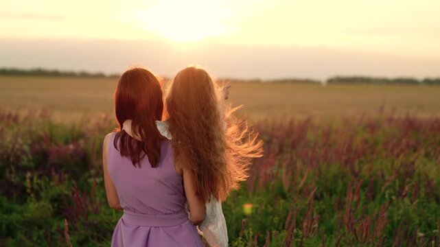 Mother carries little daughter pointing forward in country field at sunset. Happy daughter hugs mother spending vacation in park field. Mother embraces daughter in field on tranquil summer weekend