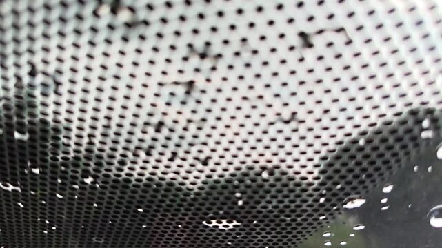 Abstract close-up view through a perforated dark screen, adorned with numerous translucent water droplets, creating an intricate, textured, and blurred perspective