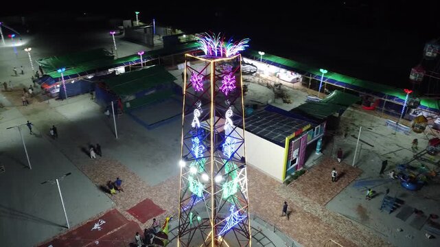 Close-up aerial view of a tall illuminated decorative tower at a night fairground with glowing led patterns and people walking in the park area lights festival amusement carnival decoration